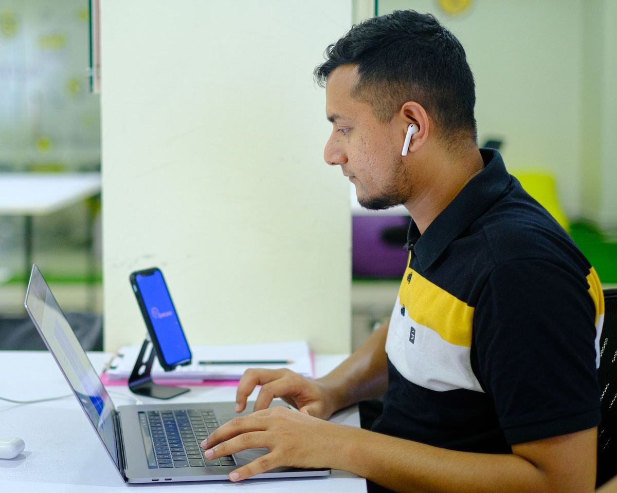 A person working on a laptop at a desk, with a phone on a stand beside it.